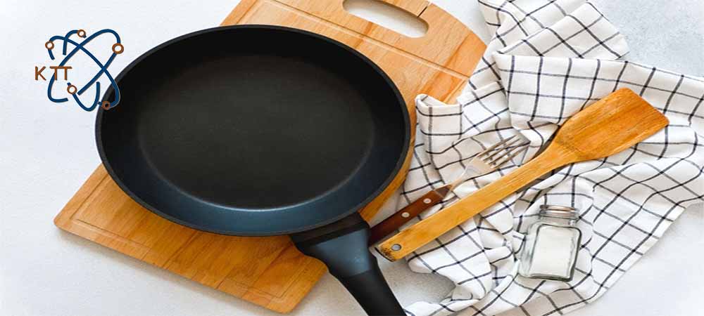 یک ماهی تابه مشکی تفلون بر روی تخته چوبی در کنار چنگال فلزی و کفگیر چوبی و نمکدان شیشه ای بر روی پارچه سفید با چهارخانه مشکی